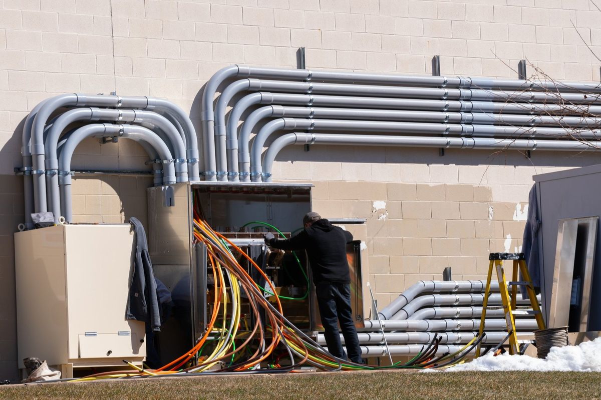 Electrician terminating feeder conductors in an outdoor switchgear cabinet beside conduit runs on a commercial building