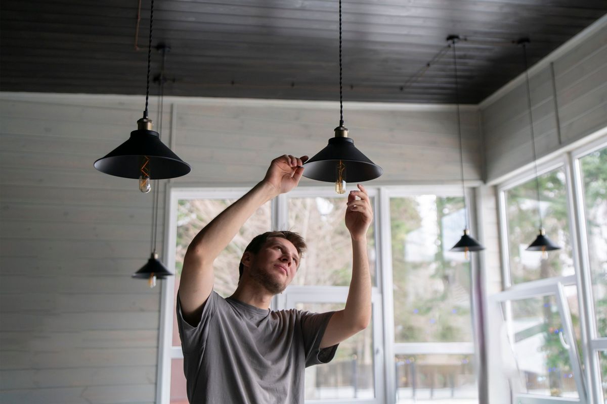 Electrician hanging black metal pendant lights with filament bulbs in a bright room with tall windows