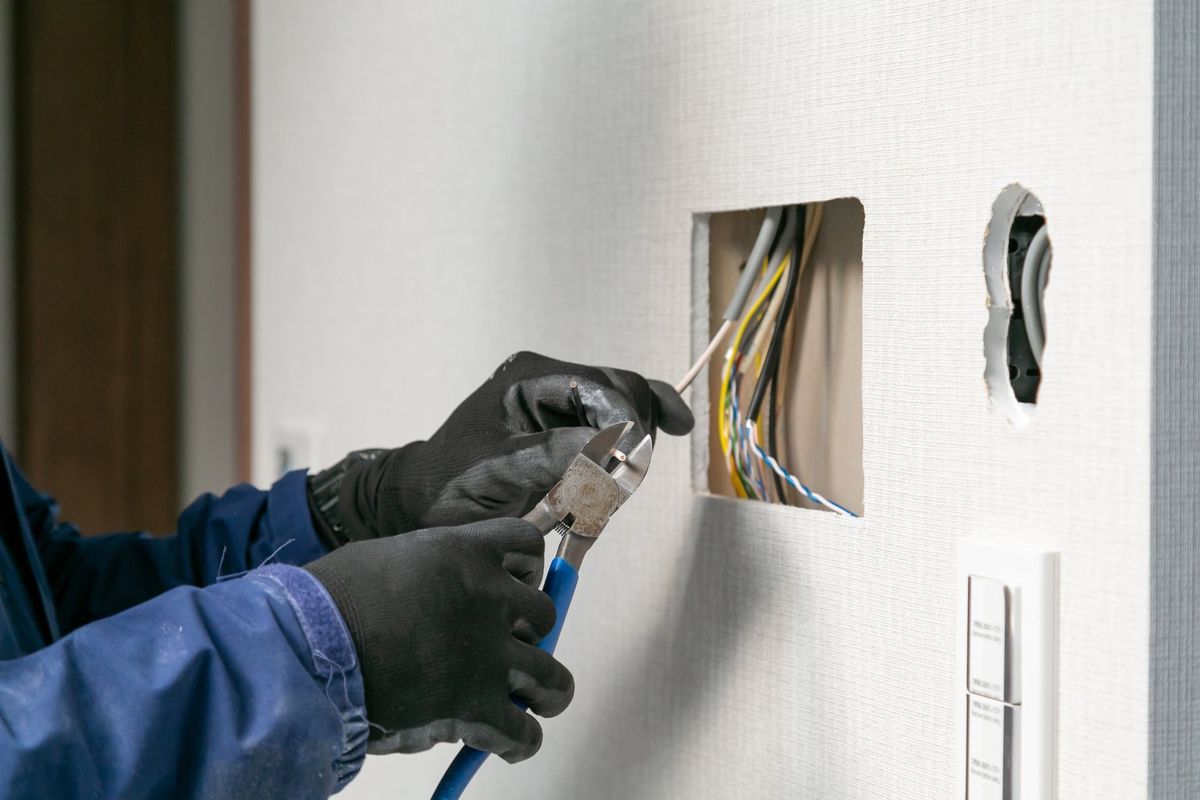 Gloved electrician trimming a cable with side cutters beside an opened wall with new wiring pulled in
